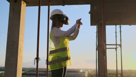 Eine-Inspektorin-Mit-VR-Brille-Und-Helm-überprüft-Den-Baufortschritt-Eines-Wolkenkratzers,-Indem-Sie-Bei-Sonnenuntergang-Ihre-Hände-Bewegt-Und-Den-Plan-Des-Gebäudes-Visualisiert